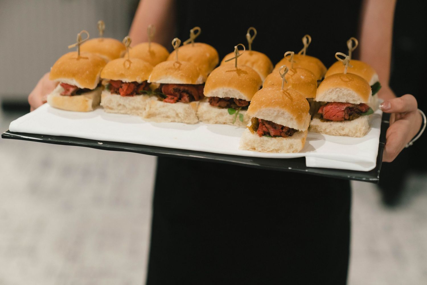 A tray of mini sliders with golden brioche buns being served at a catering event on a white platter.