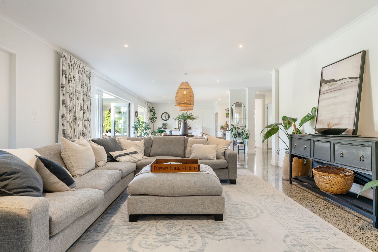 Spacious living area with grey sectional, ottoman and rattan pendant light overlooking garden views.