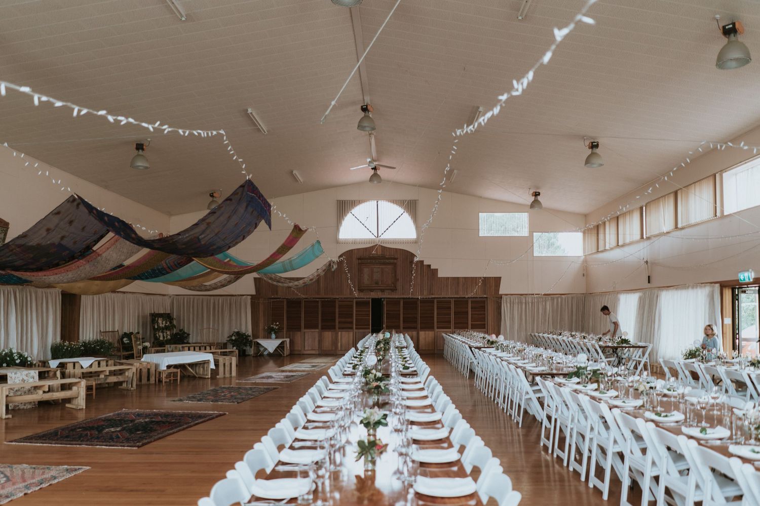 An elegant wedding reception hall with long dining tables, white chairs, and draped fabric decorations across the ceiling.