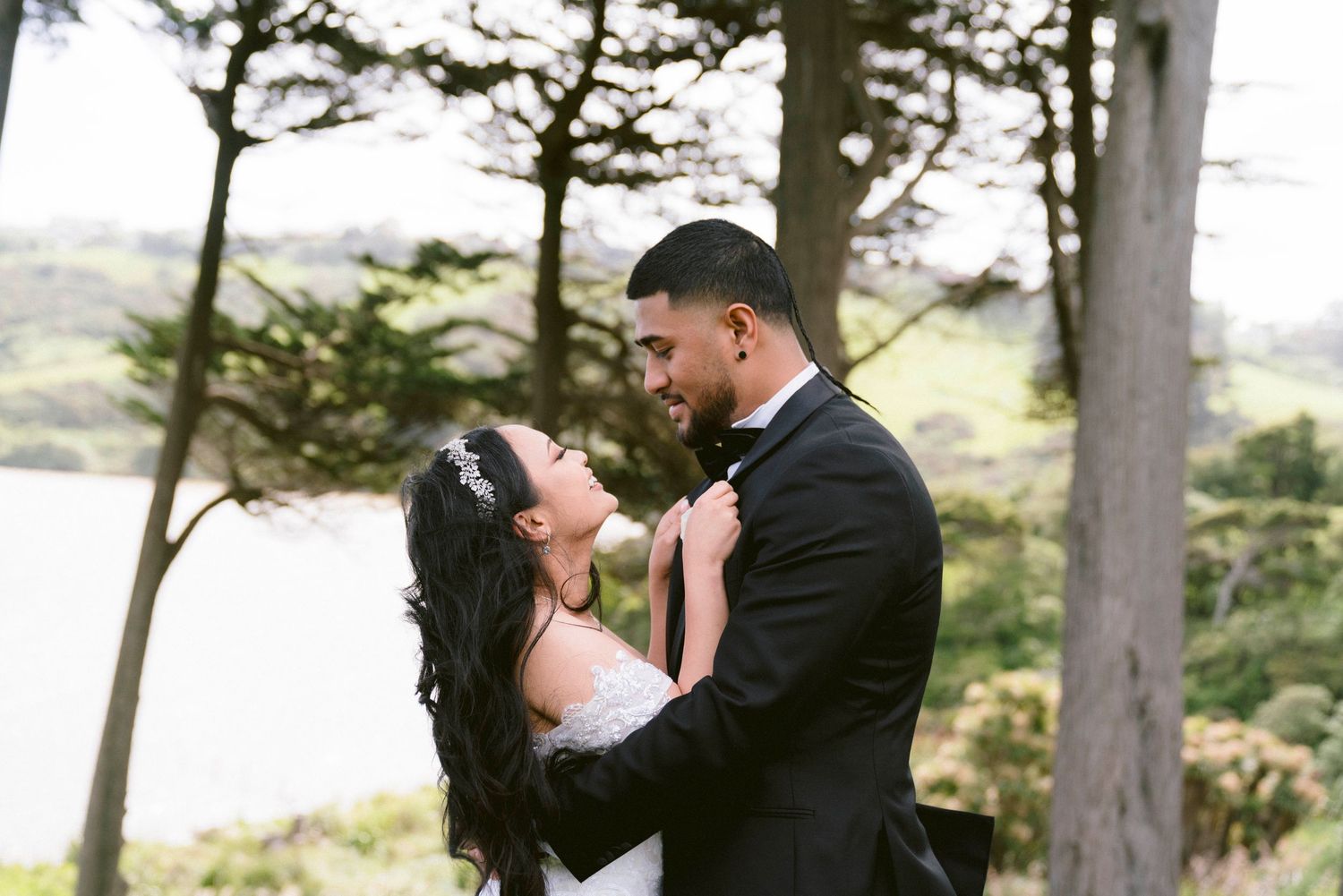 A romantic couple embraces outdoors with trees and water in the background during their wedding photoshoot.