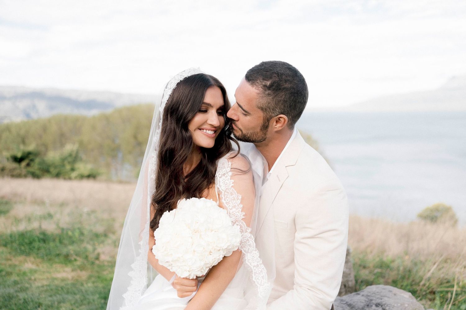 Newlywed couple shares intimate moment on scenic coastal cliff with white floral bouquet and flowing veil.