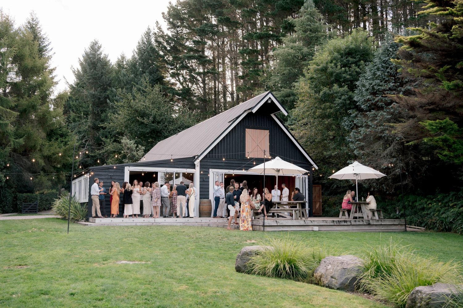 A rustic black barn venue surrounded by pine trees hosts wedding guests on its lawn.