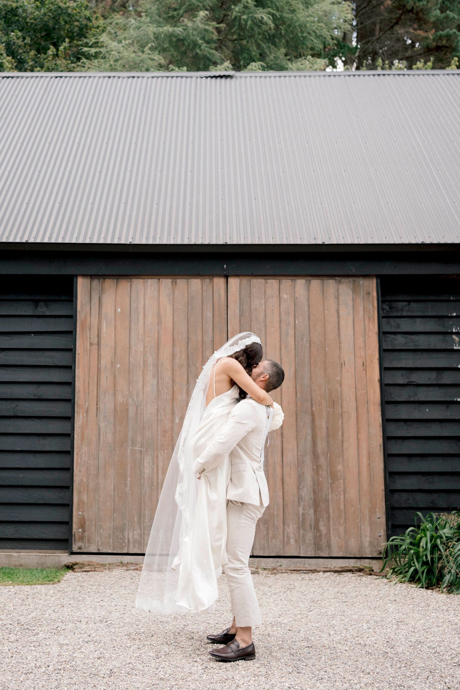 Romantic wedding couple embrace in front of rustic barn with metal roof.
