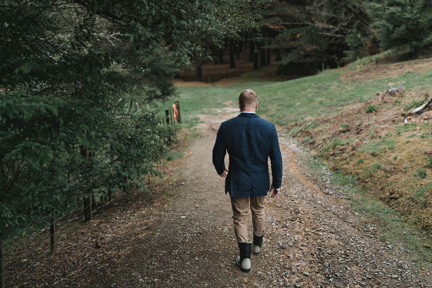 A person in casual attire walking down a scenic forest path surrounded by evergreen trees and sloping terrain.