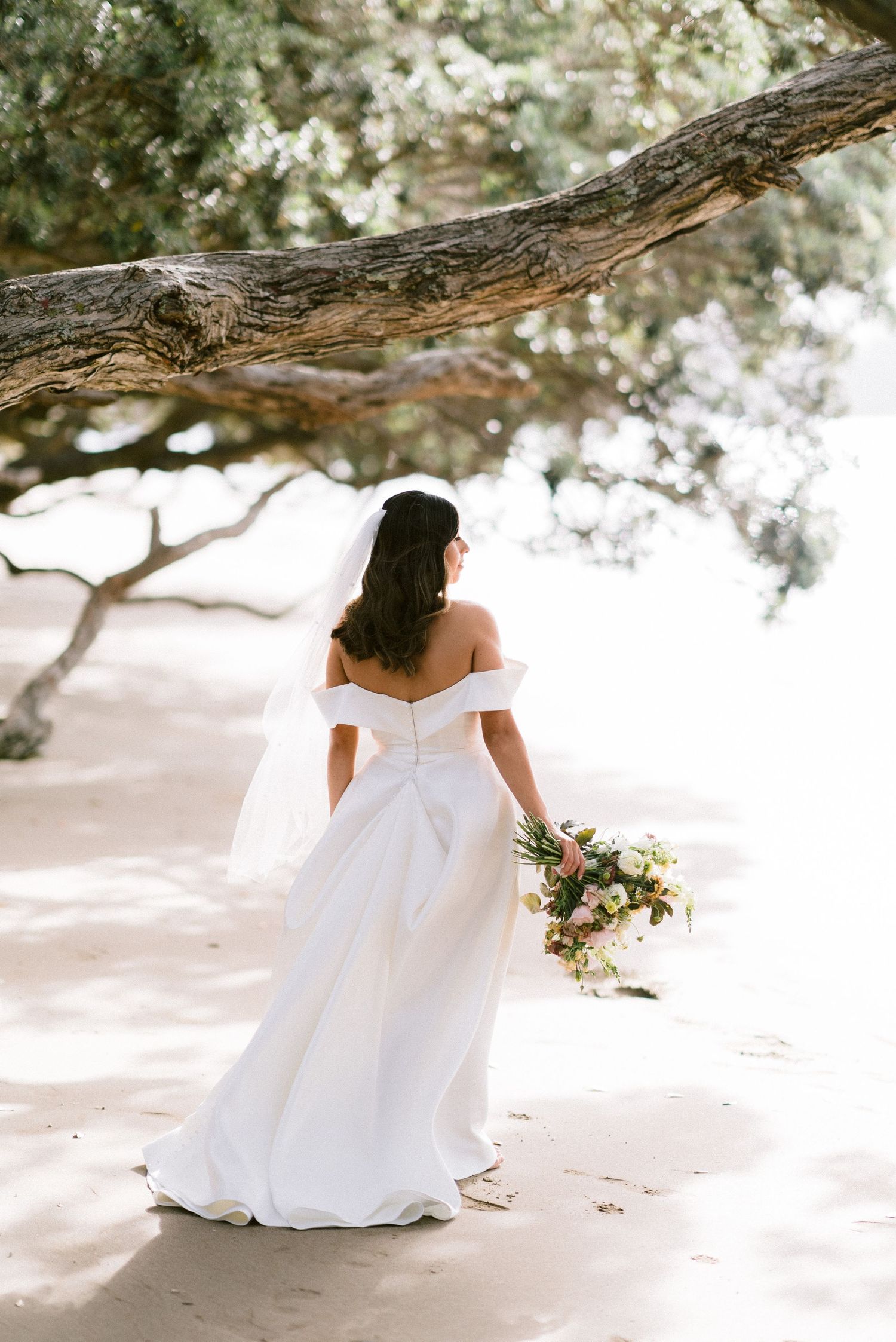 Elegant bride in off-shoulder dress holds bouquet while standing under flowering tree branches.