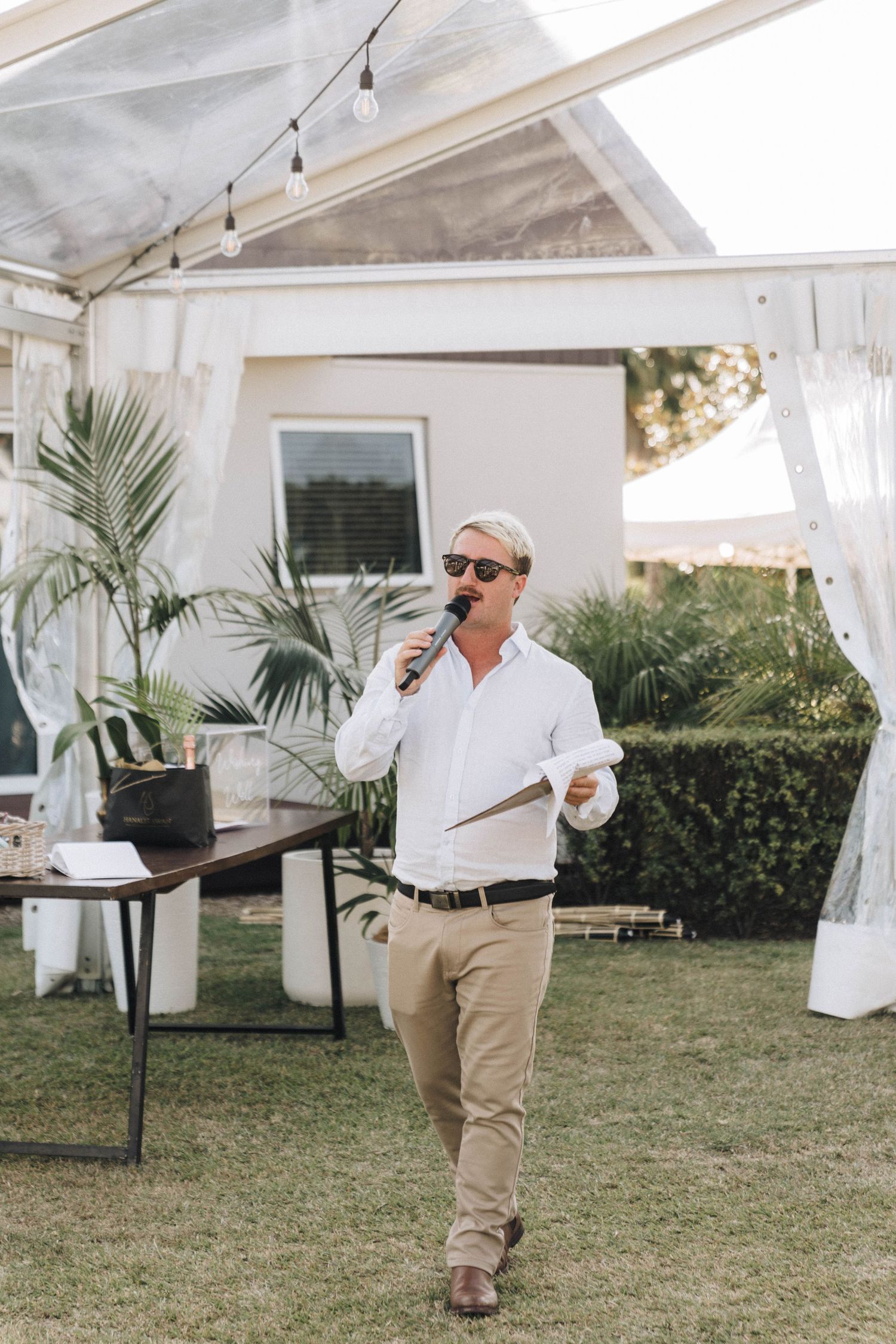 Someone in a white button-down shirt and khaki pants speaks into a microphone under an outdoor tent at a special event.