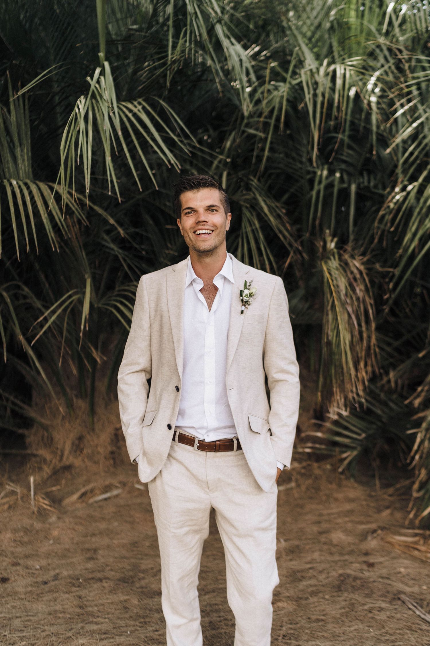 A groom in a light beige linen suit and white shirt poses in front of tropical palm fronds.