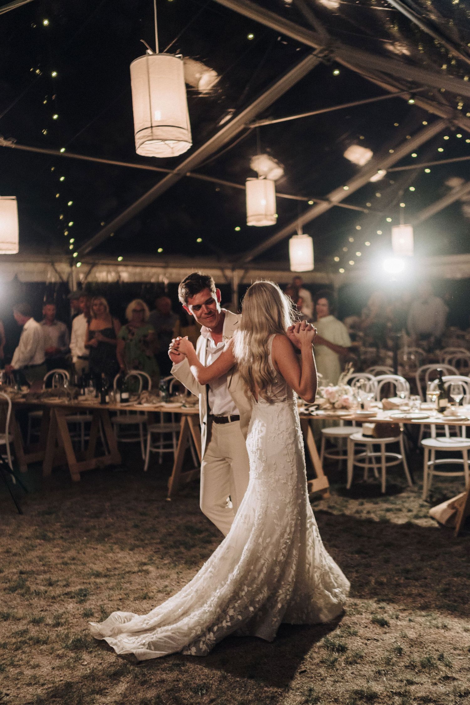 Romantic wedding couple dancing at an evening reception under illuminated tent with fairy lights.