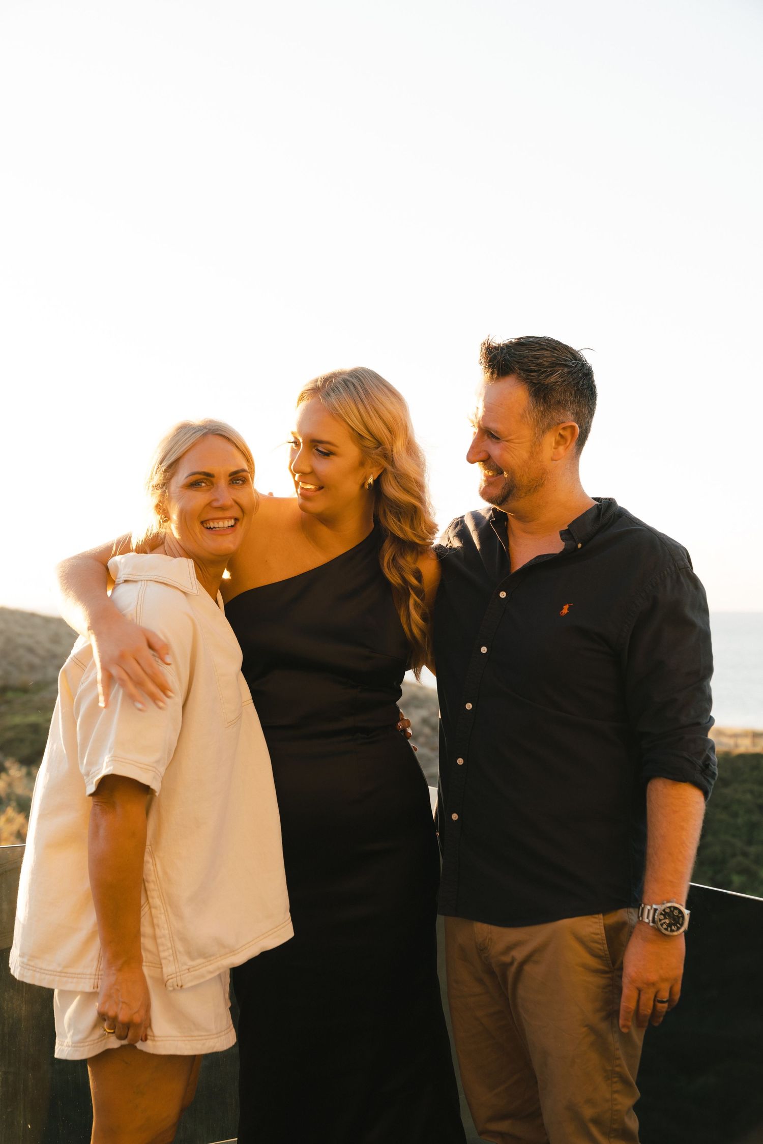 Three friends share a joyful moment together at sunset, with one wearing black and others in casual attire.