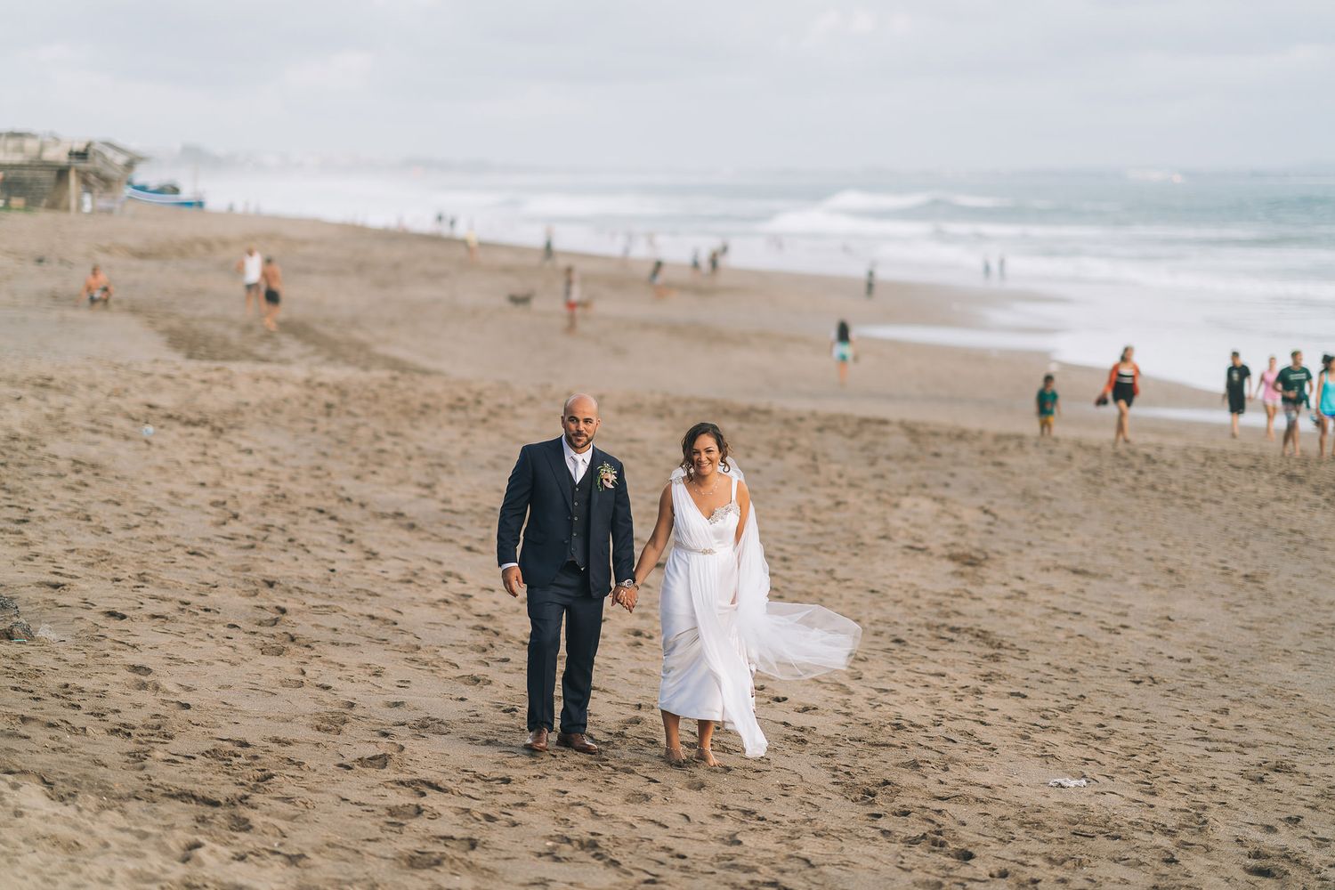 A couple walks hand in hand along a misty beach shoreline with other beachgoers visible in the background.