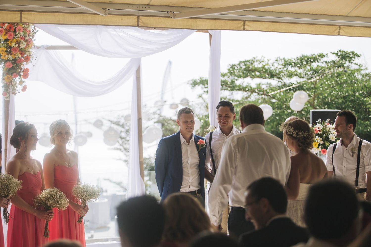 Wedding ceremony under a white draped canopy with floral decorations and guests gathered outdoors.