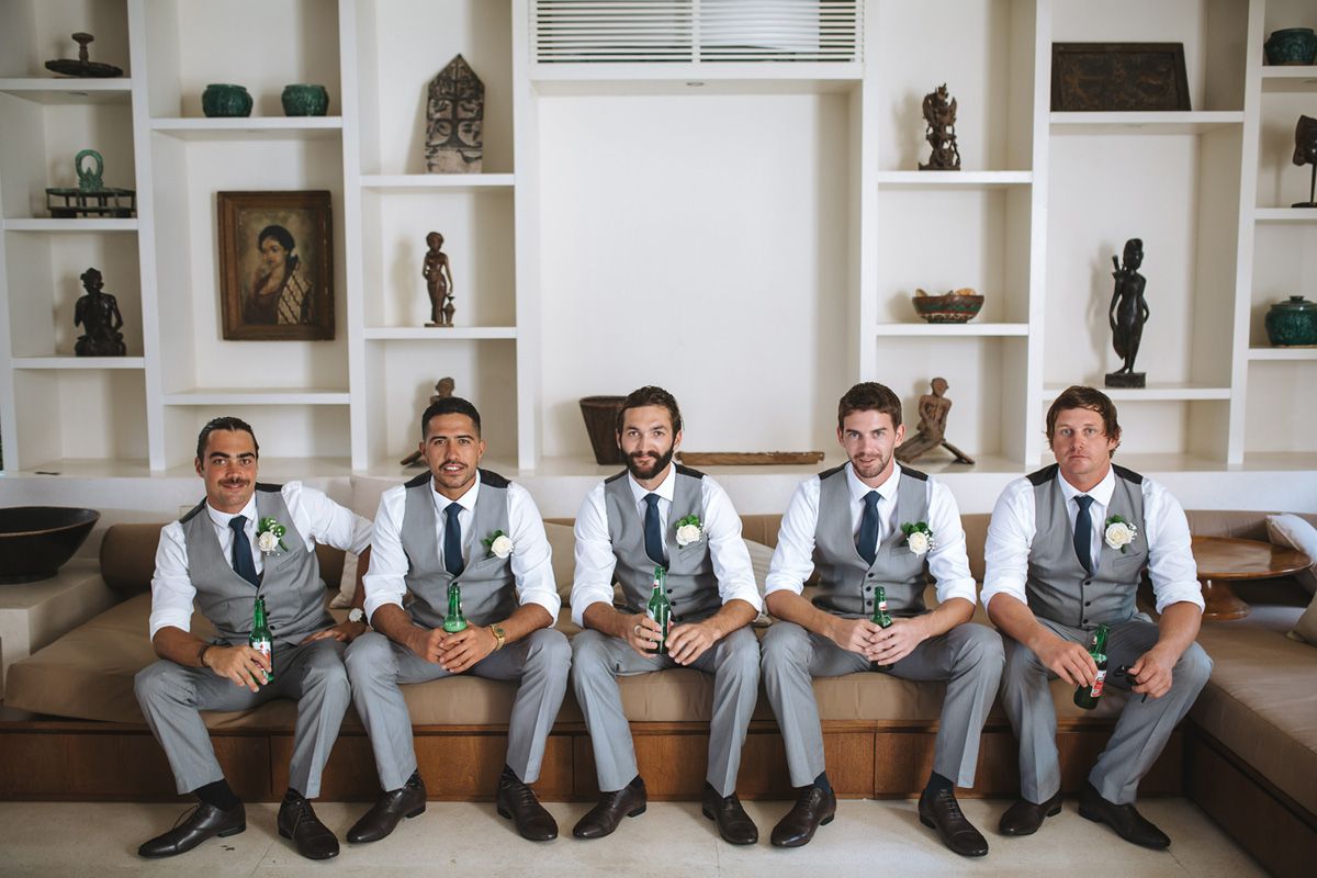 Five groomsmen in matching gray suits and blue ties sit together on a white built-in bench with decorative shelving behind them.
