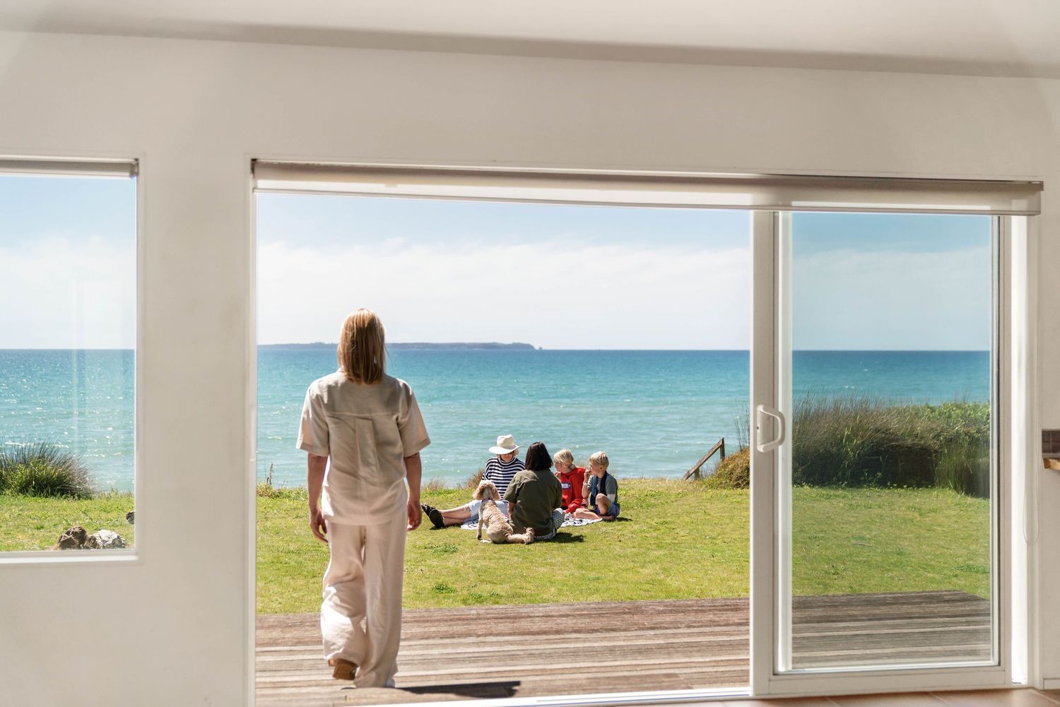 Five panoramic windows overlooking a beach with window shades in various positions frame a tranquil ocean view.