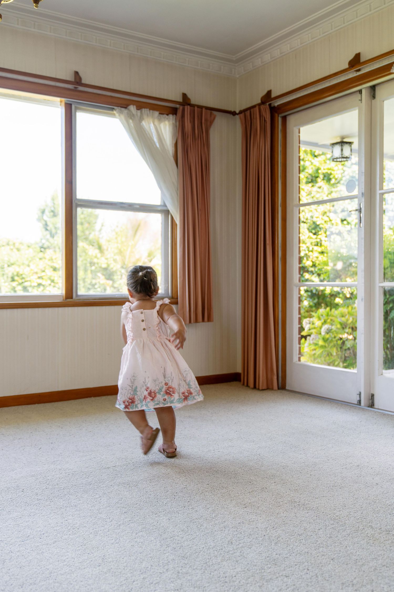 An unfurnished carpeted room with white walls features tall windows, wooden trim, and sheer curtains in natural daylight.