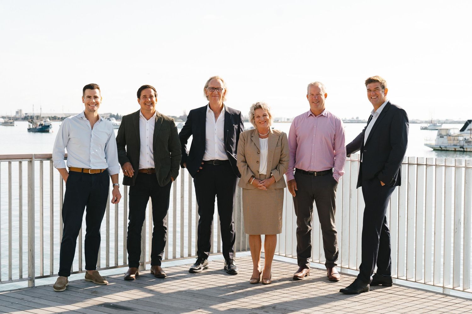 Wedding party members lined up on a bright waterfront terrace with boats and harbor views in the distance.