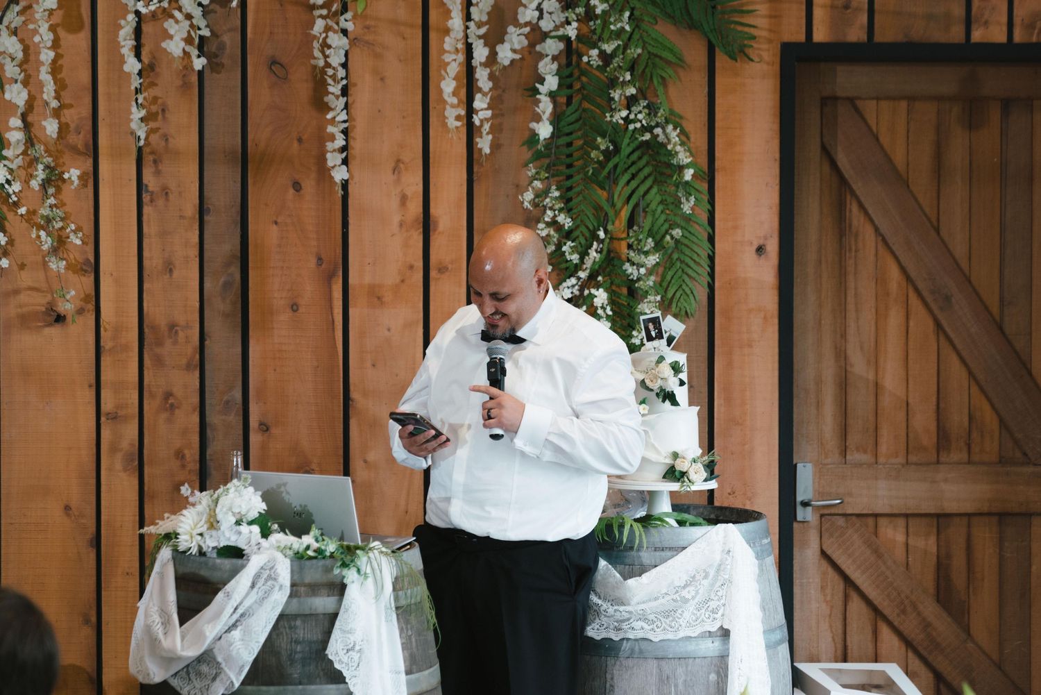 Speaker gives a wedding toast at rustic venue with wooden walls decorated with floral garlands.
