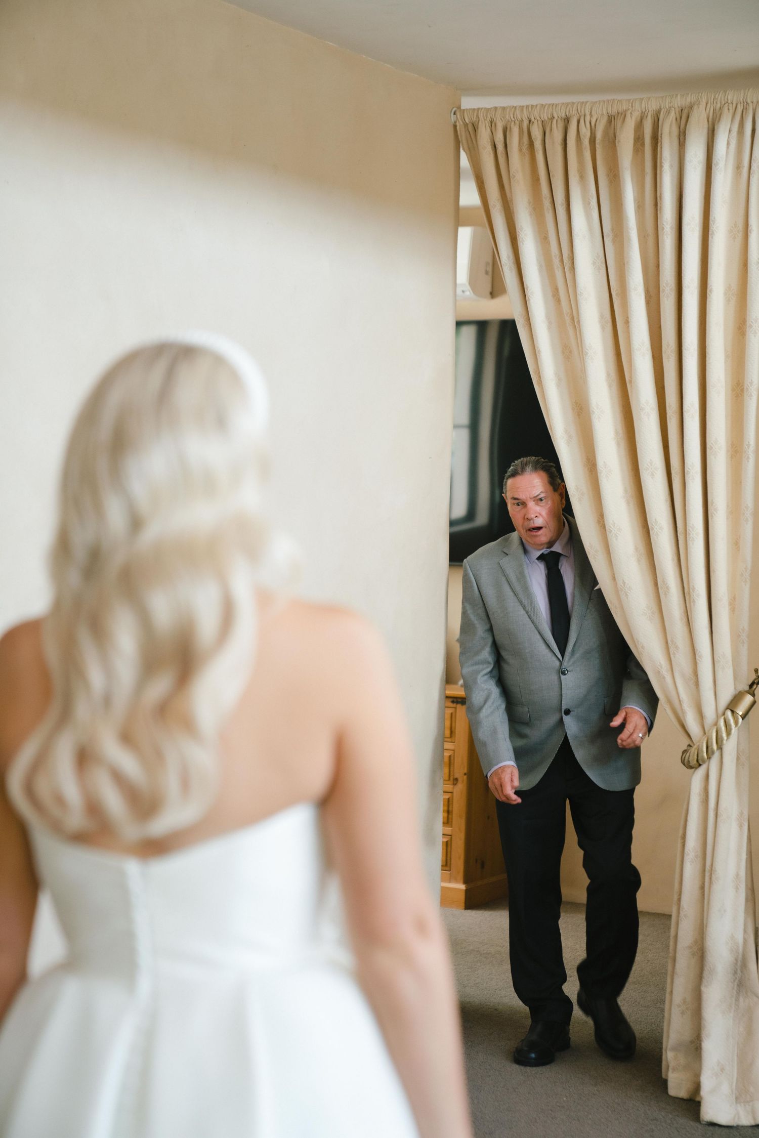 An emotional first look between a bride in a white gown and someone peeking through cream-colored curtains.