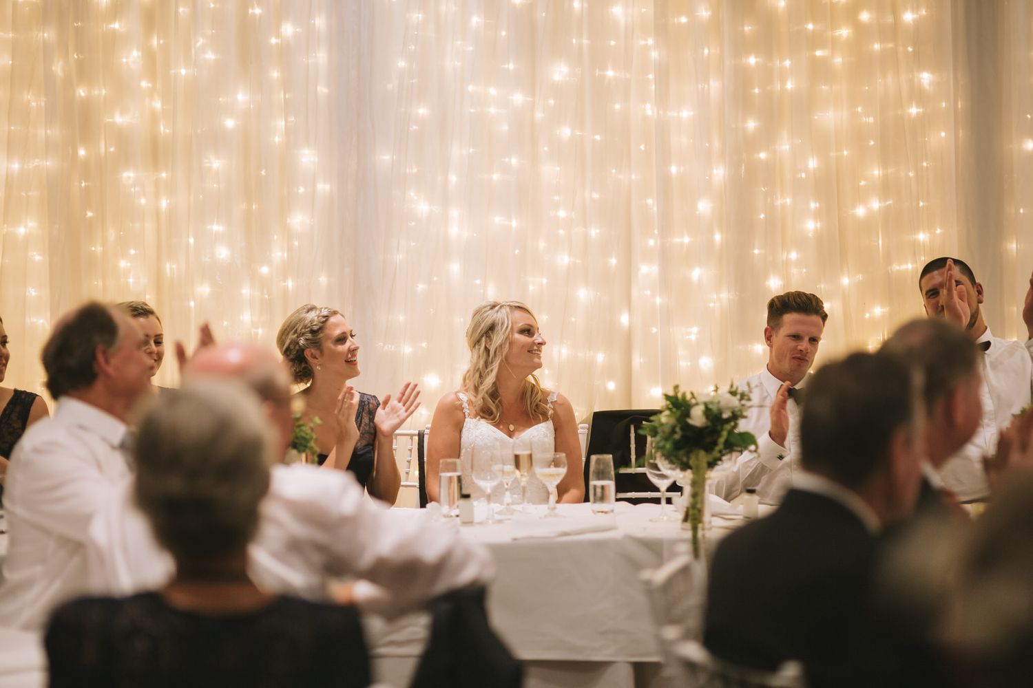 Wedding reception guests applaud during speeches at an elegant venue with sparkling string lights in the background.