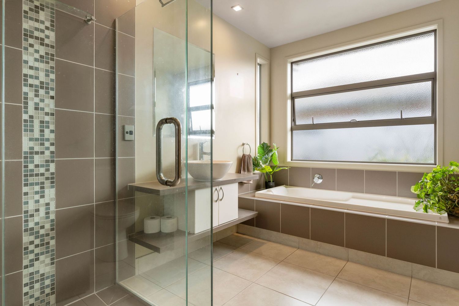 Modern bathroom with glass shower enclosure, taupe tile walls, mosaic accent strip and large frosted window.