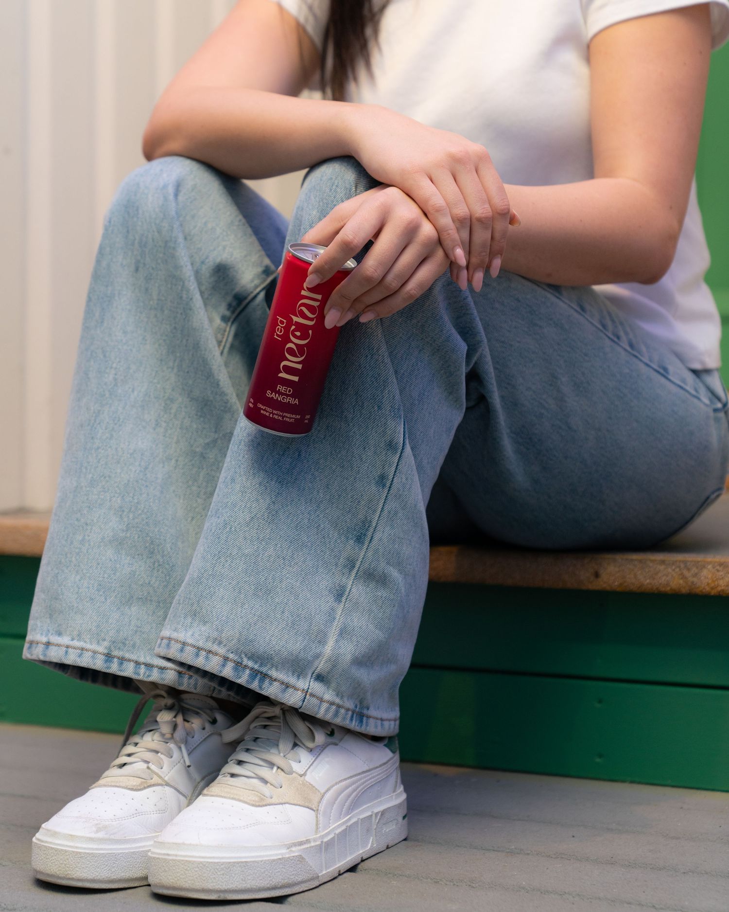 A casual shot of someone in jeans and white sneakers holding a red Nectar drink can.