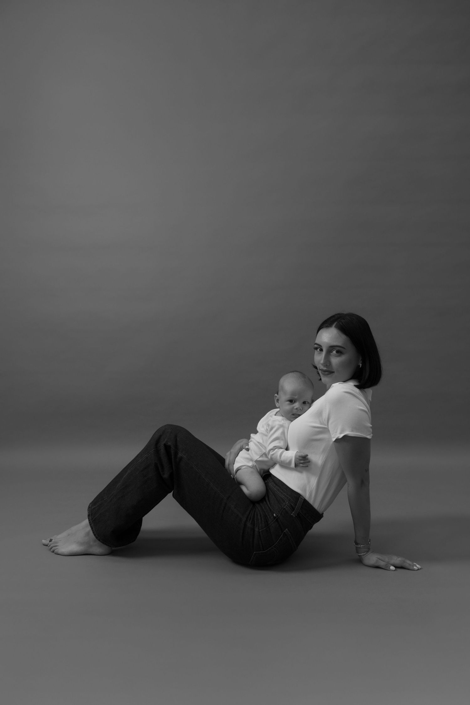 Black and white photo of a young mother sitting on floor holding her newborn baby, wearing white t-shirt and jeans.