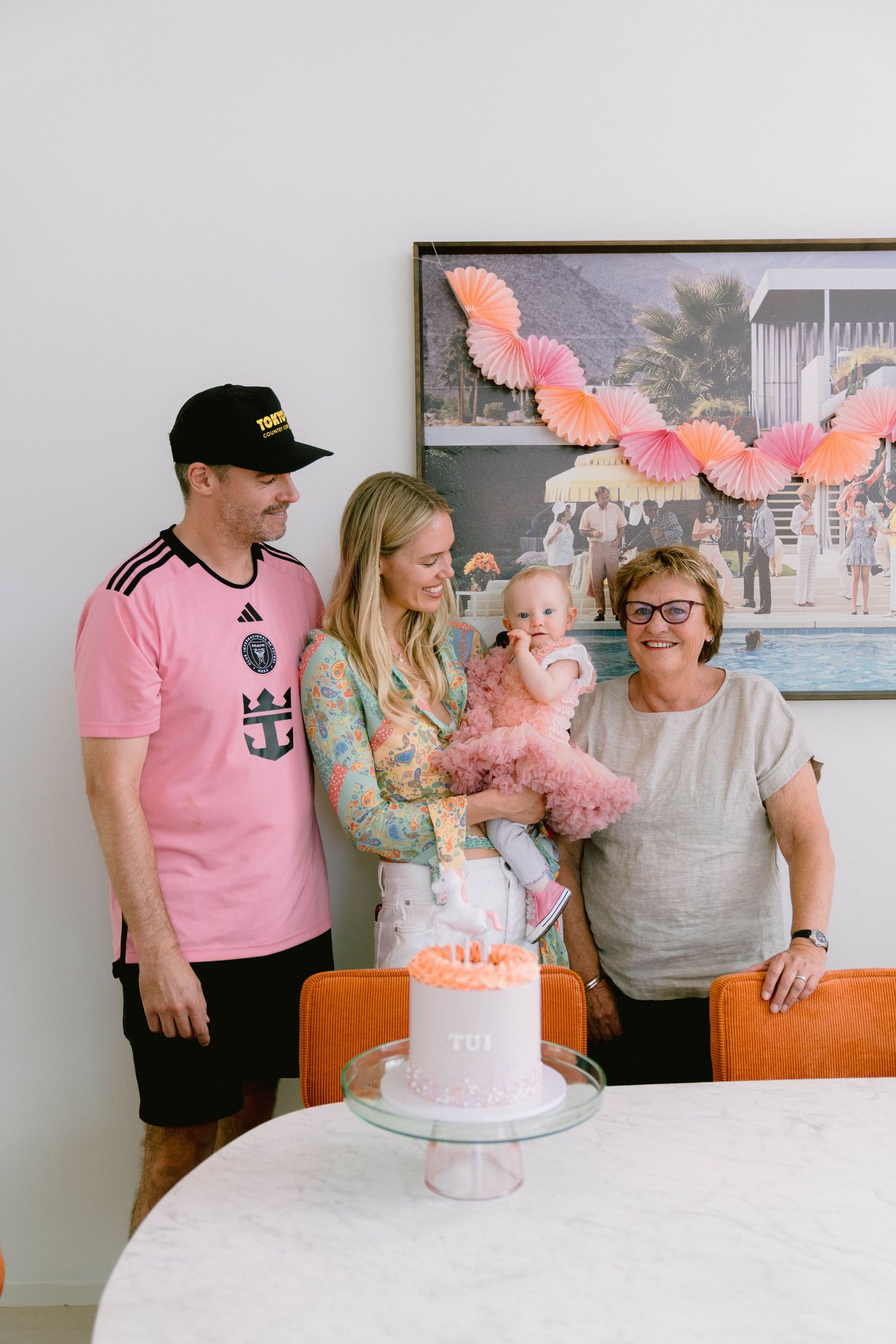A pink birthday celebration with a white cake on a table, decorated with coral paper fans hanging on the wall behind.