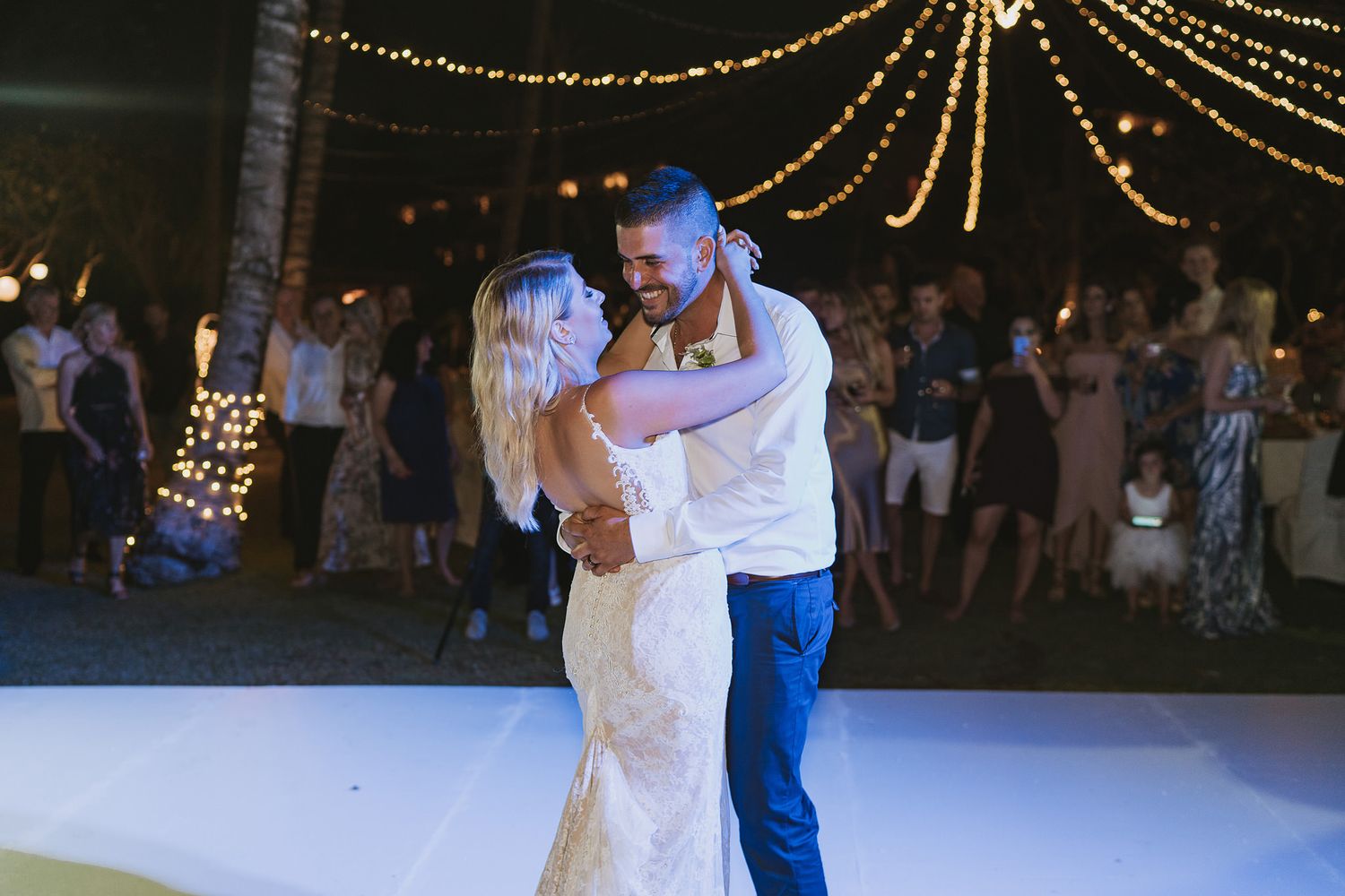 Romantic first dance at night under string light canopy at outdoor wedding reception.