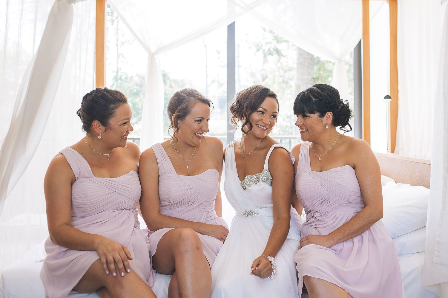 Four bridesmaids in matching lavender dresses share joyful candid moments together during wedding preparations.