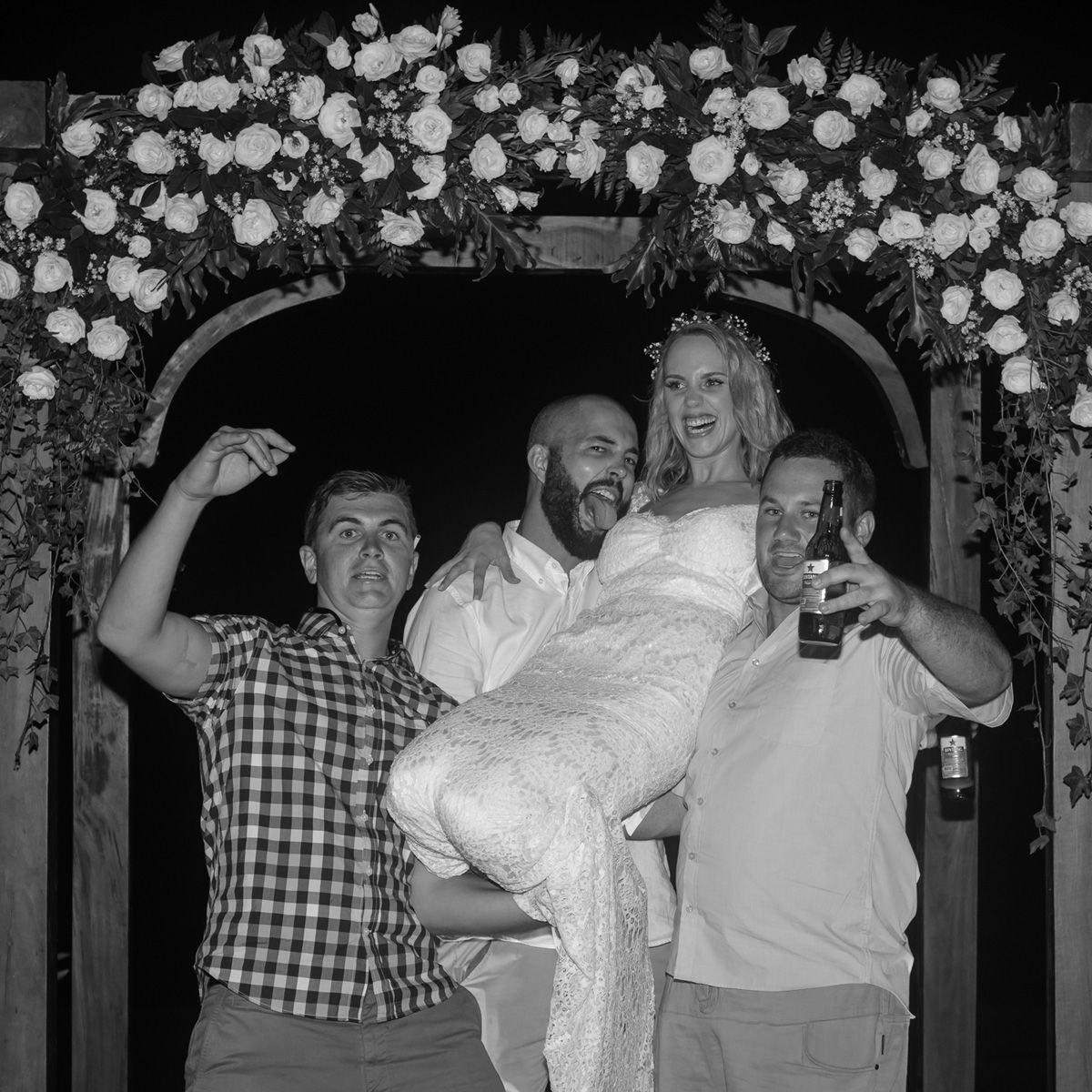 A black and white photo of a celebration under a floral arch with a joyful group posing and having fun.
