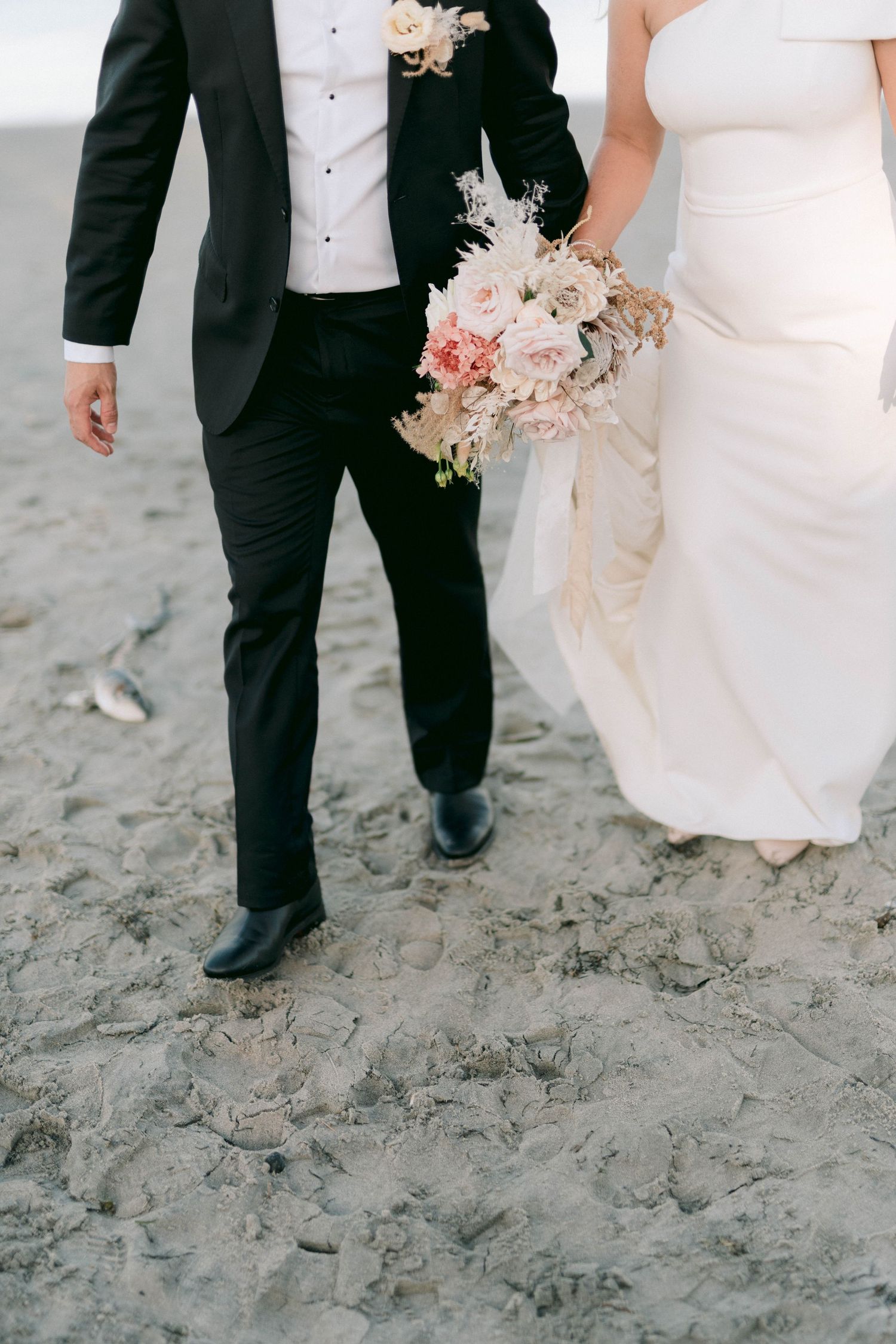 Close-up of wedding party feet walking on beach sand showing formal shoes and white dresses.