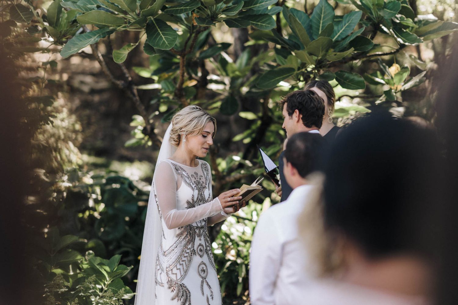 Two people in white dresses share an intimate moment under green garden foliage during a wedding ceremony.