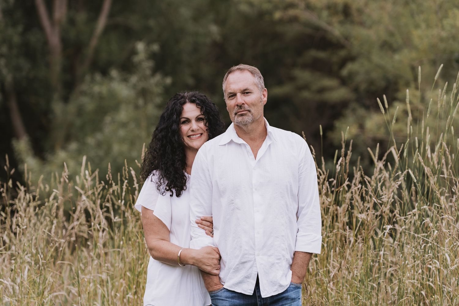 A couple wearing white clothing embraces while standing in tall grass and wildflowers in a natural outdoor setting.