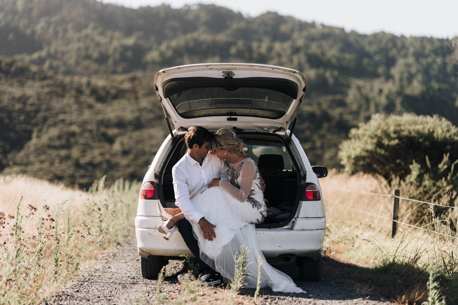 Two casually dressed people sit in SUV trunk on mountain road with scenic hillside view in background.