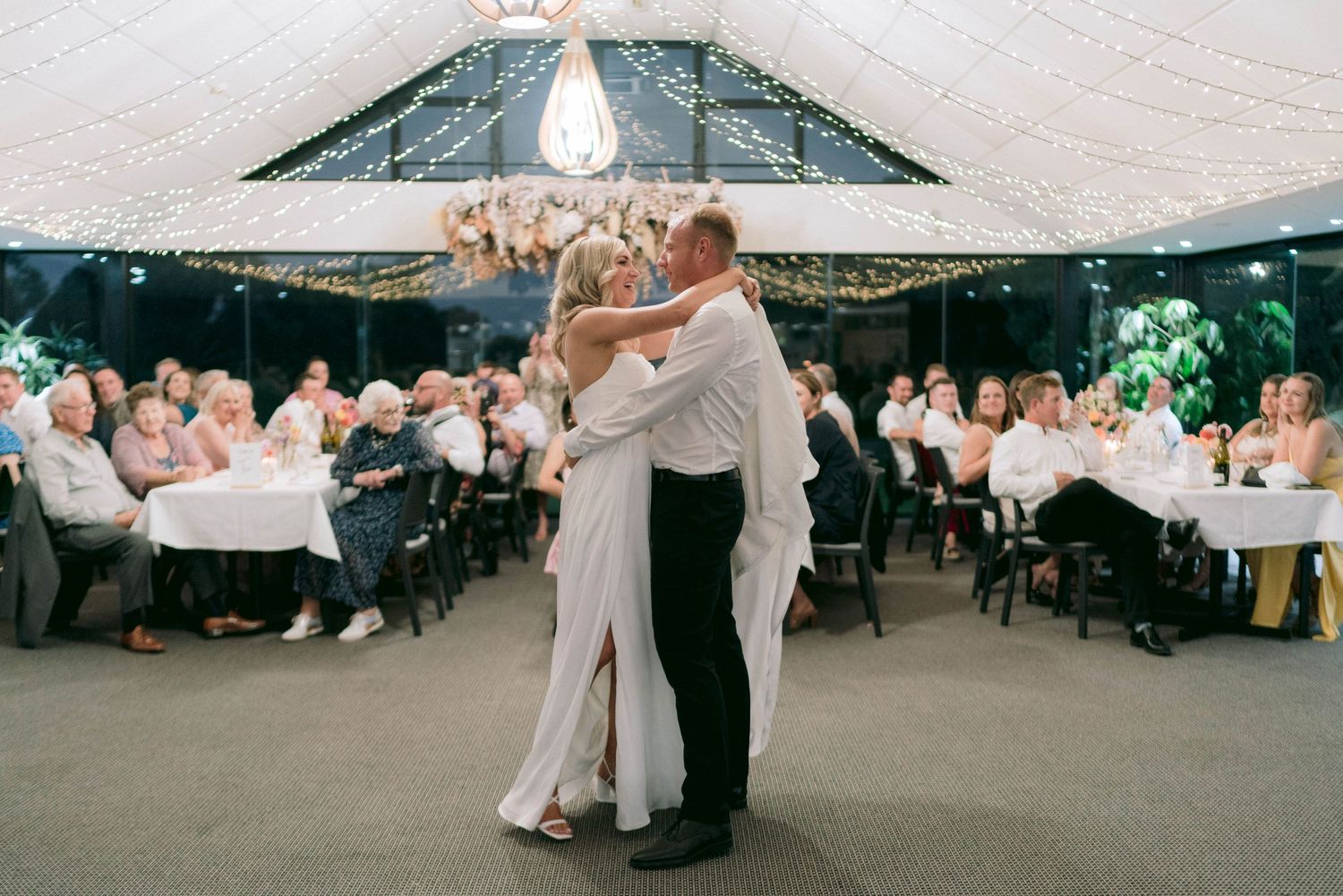 Couple shares their first dance under tent with string lights while wedding guests look on from tables.