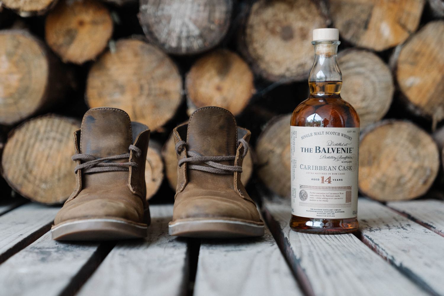 A bottle of Balvenie whisky sits beside brown leather boots on weathered wooden planks with stacked logs in background.