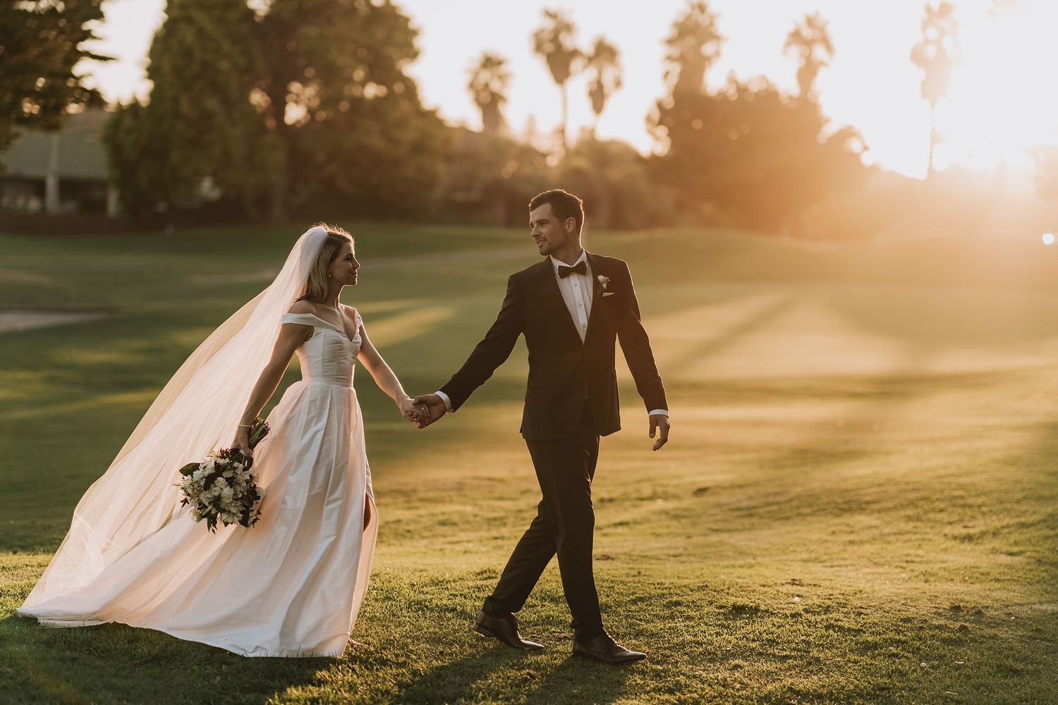 Newlywed couple walking hand in hand across sunlit golf course during golden hour with trailing white veil and bouquet.