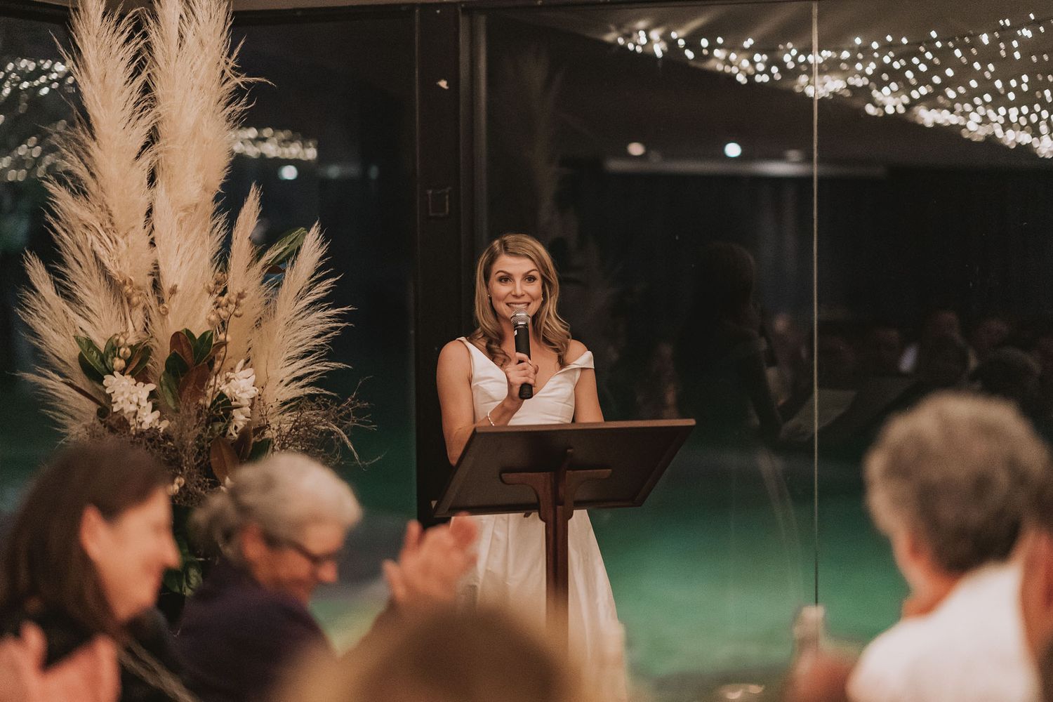 Wedding reception speech given at rustic venue decorated with pampas grass and string lights at night.