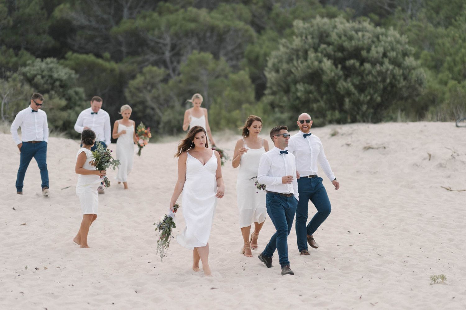 A group wearing coordinated white and blue outfits walks joyfully together on a sandy beach during a photo session.