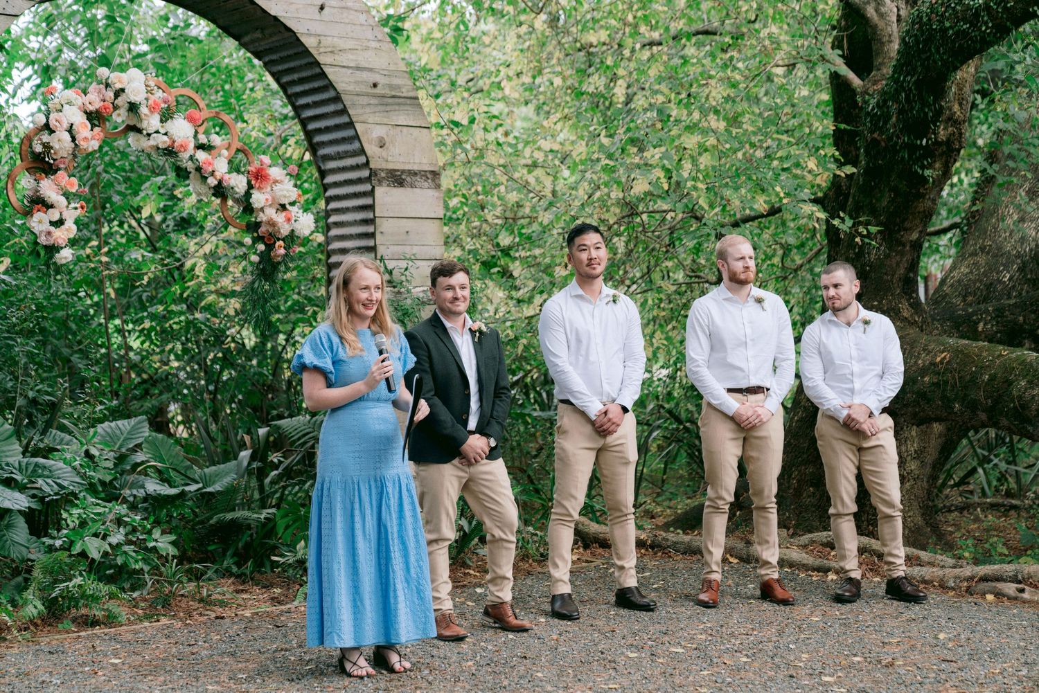 Wedding party members in light blue dresses and khaki suits stand in line against lush garden greenery.