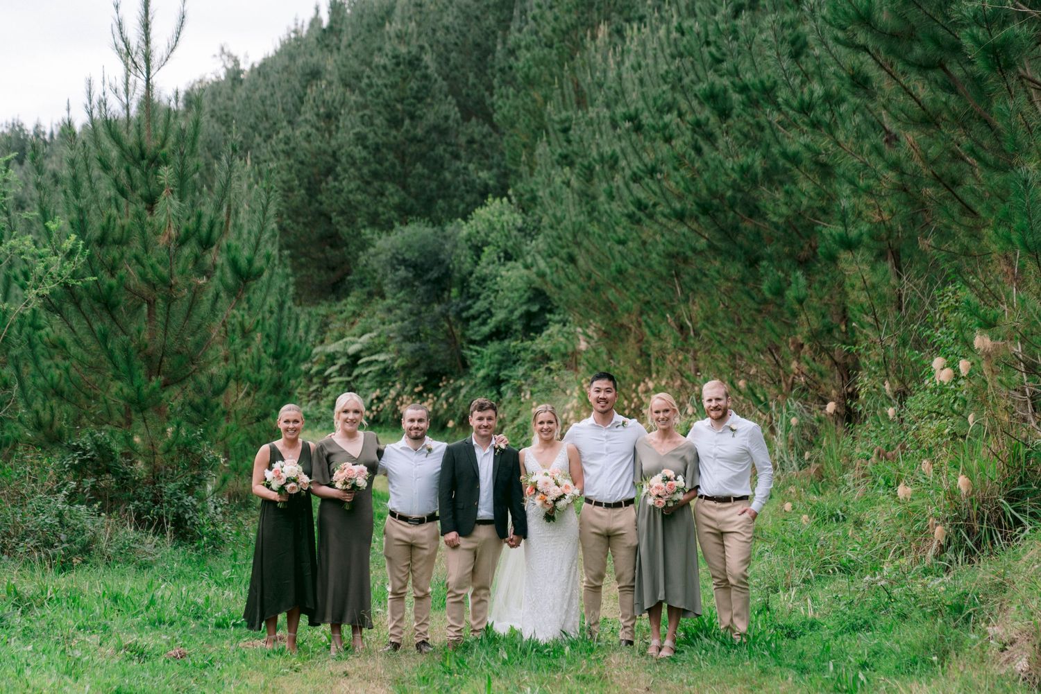 Wedding party poses for a photo in front of evergreen trees on a grassy hillside.