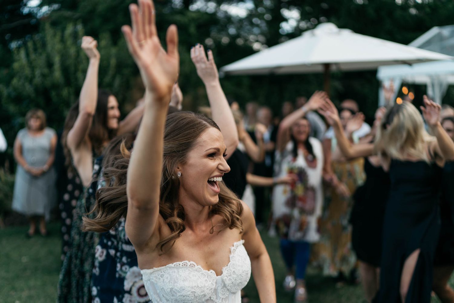 Celebrating crowd raises arms in joy at outdoor wedding reception with white tent in background.
