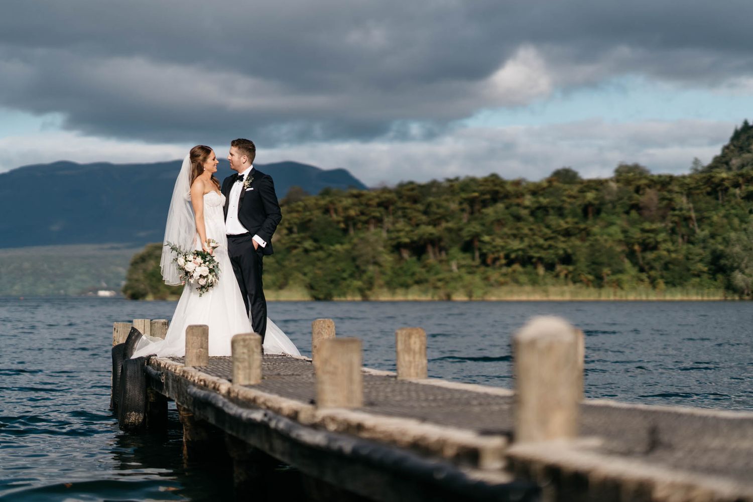 A couple in wedding attire stands together on a wooden dock overlooking a scenic mountain lake under dramatic clouds.