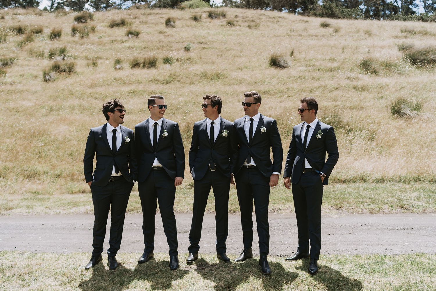 Groomsmen in black suits and ties walking along a golden hillside path during a wedding photo session.