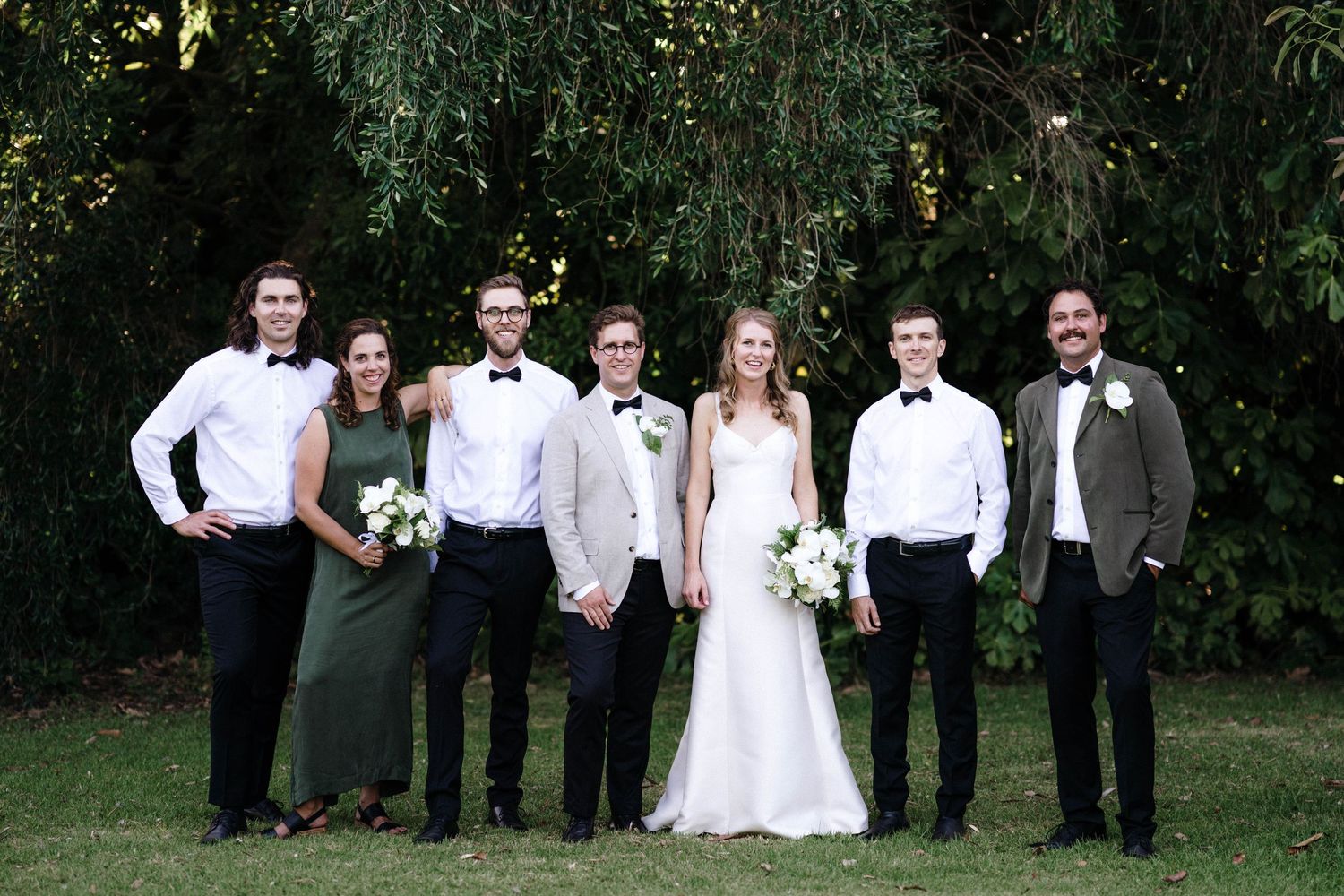 A wedding party poses together outdoors against a backdrop of lush green trees and foliage.