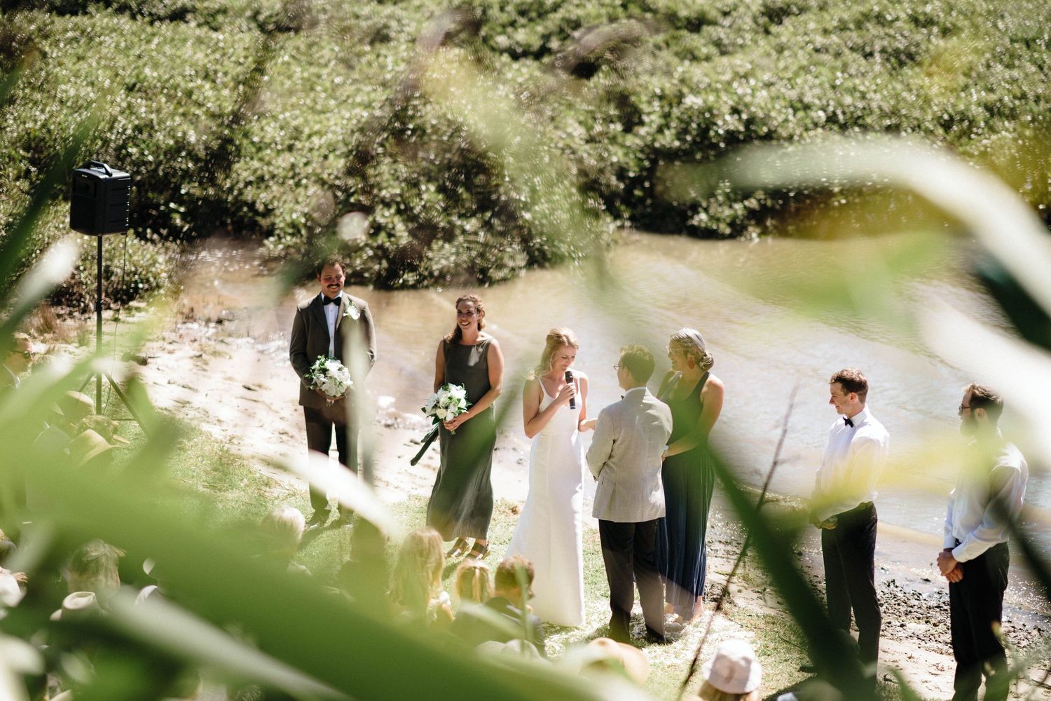 An outdoor wedding ceremony being photographed through greenery with a natural sunlit glow in the background.