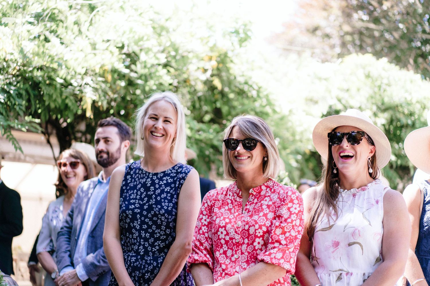 Wedding guests in colorful summer attire smile and laugh while seated outdoors during a sunny celebration.