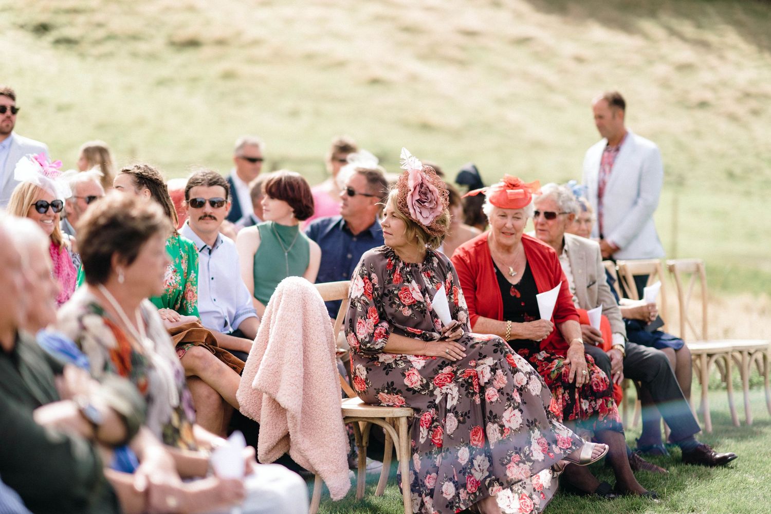 Wedding guests seated outdoors on wooden chairs during a sunny ceremony.
