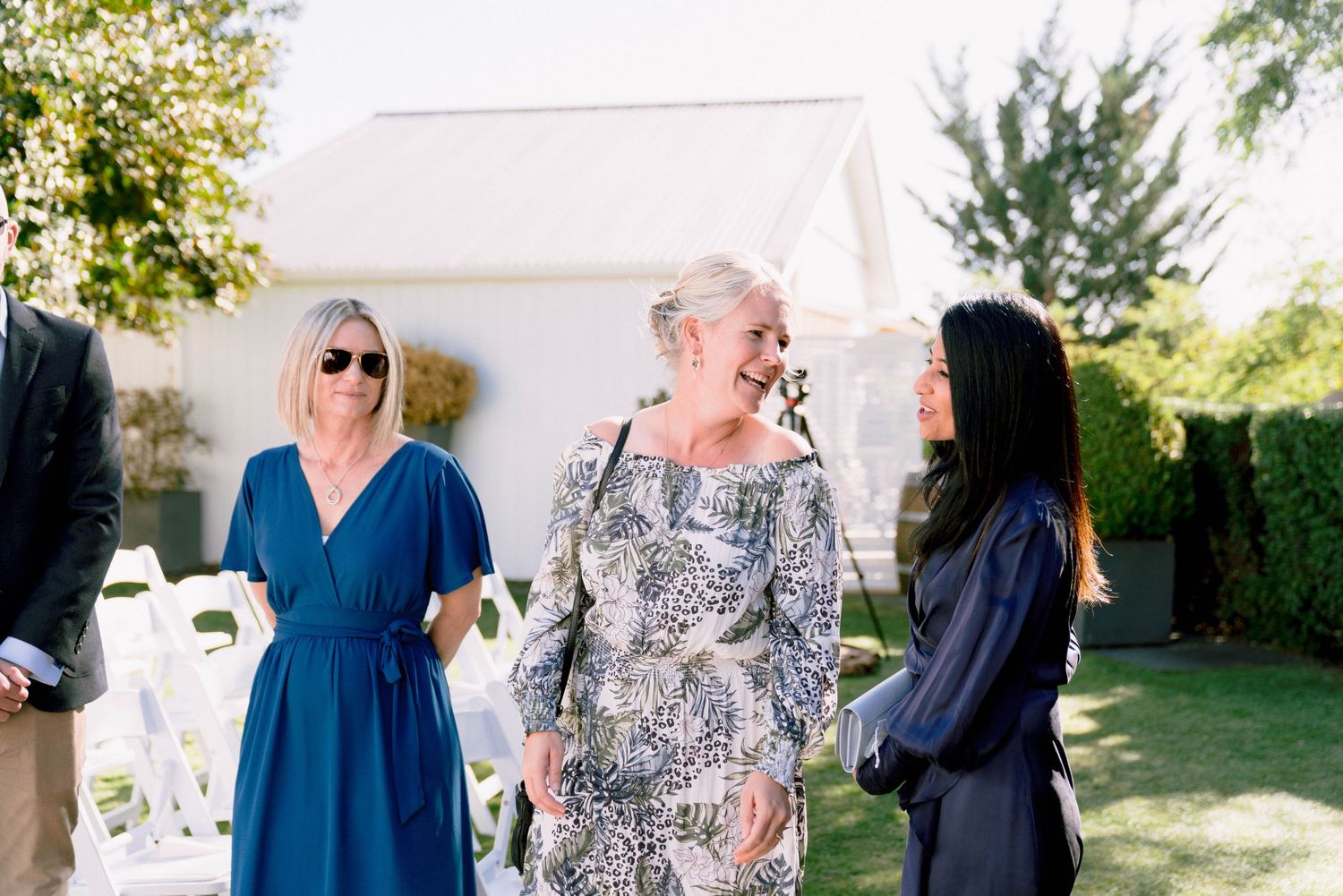 Three people engaged in conversation at an outdoor gathering with sunlit greenery in the background.