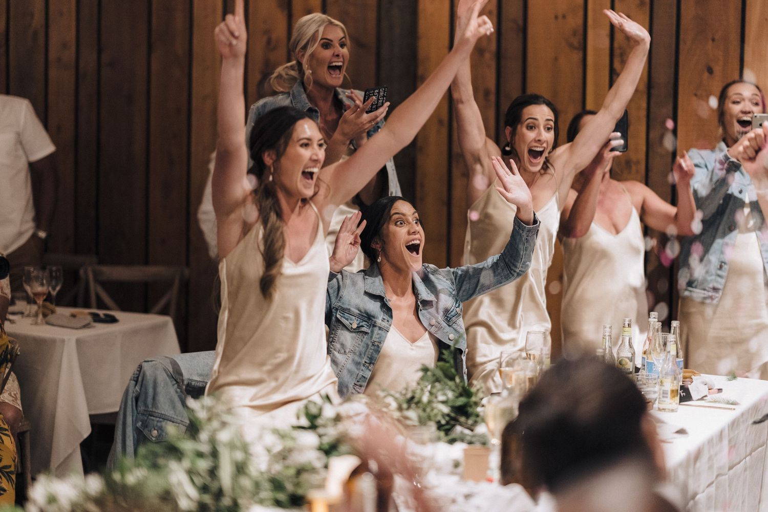 Wedding guests celebrate enthusiastically with raised arms at a rustic indoor reception venue.