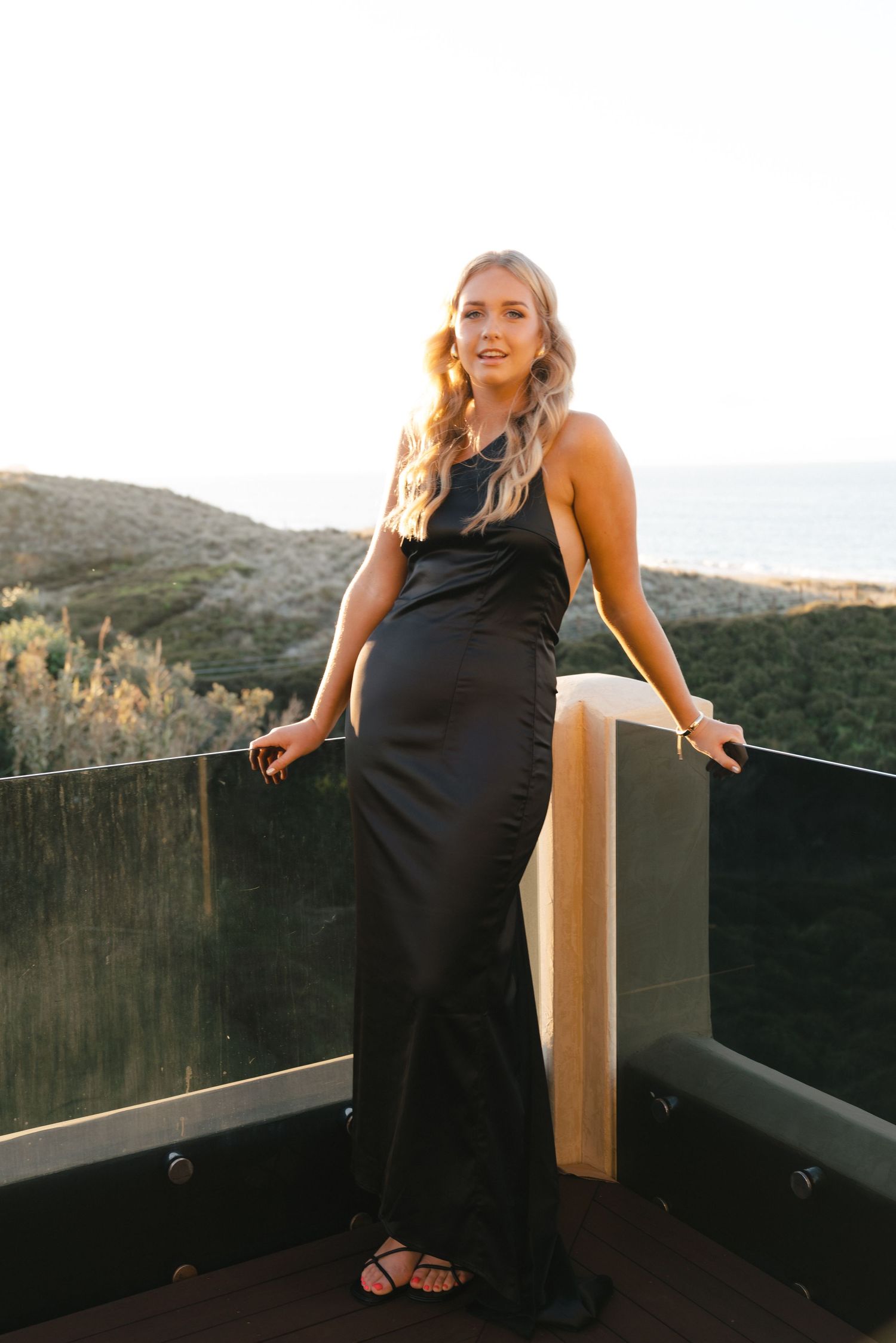 A person in a black formal dress poses on a deck overlooking scenic mountain views at sunset.